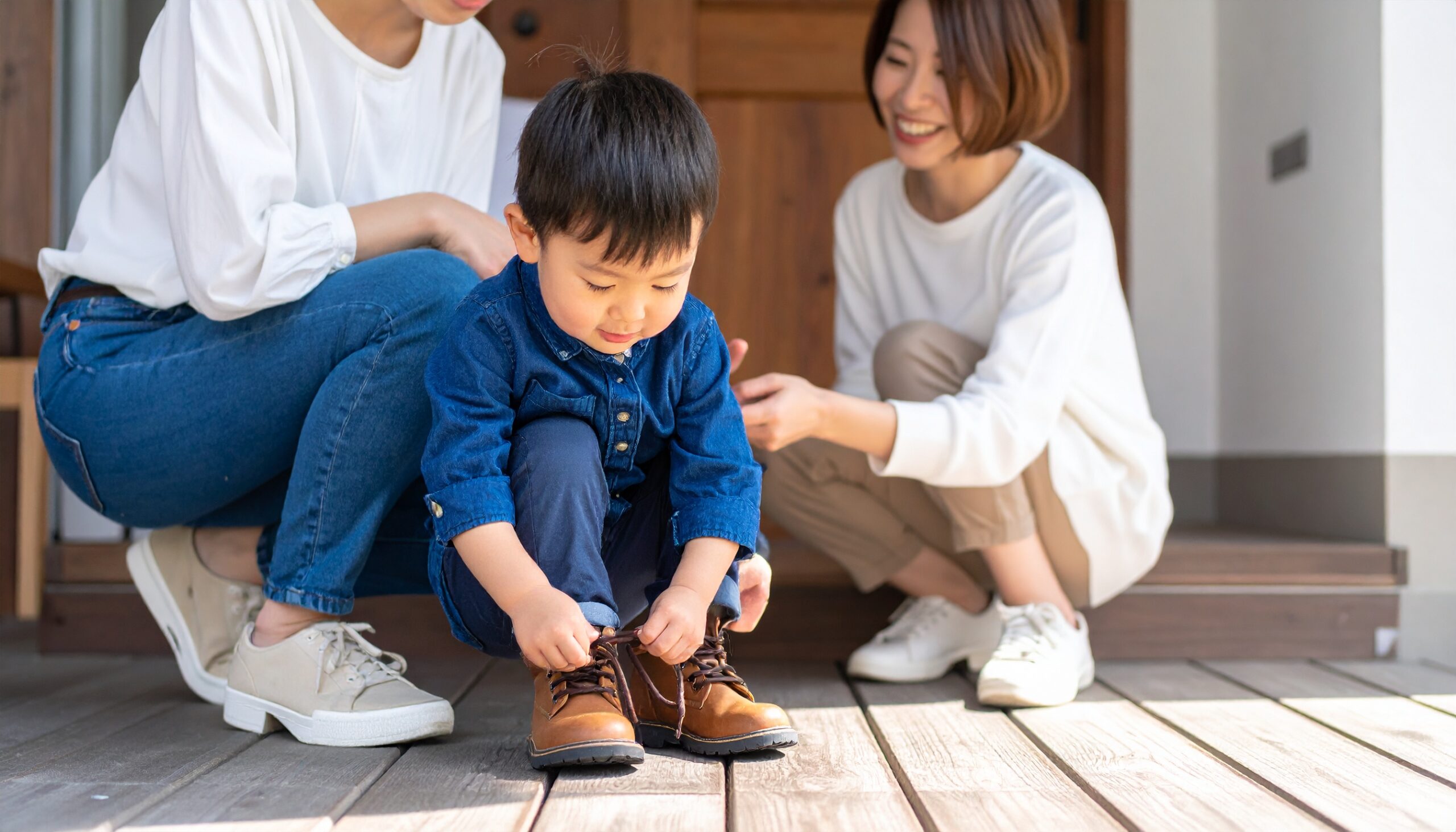 <img src="画像URL" alt="3歳の日本人の男の子が自分で靴を履こうとしている姿を、母親が見守っている様子。" />
