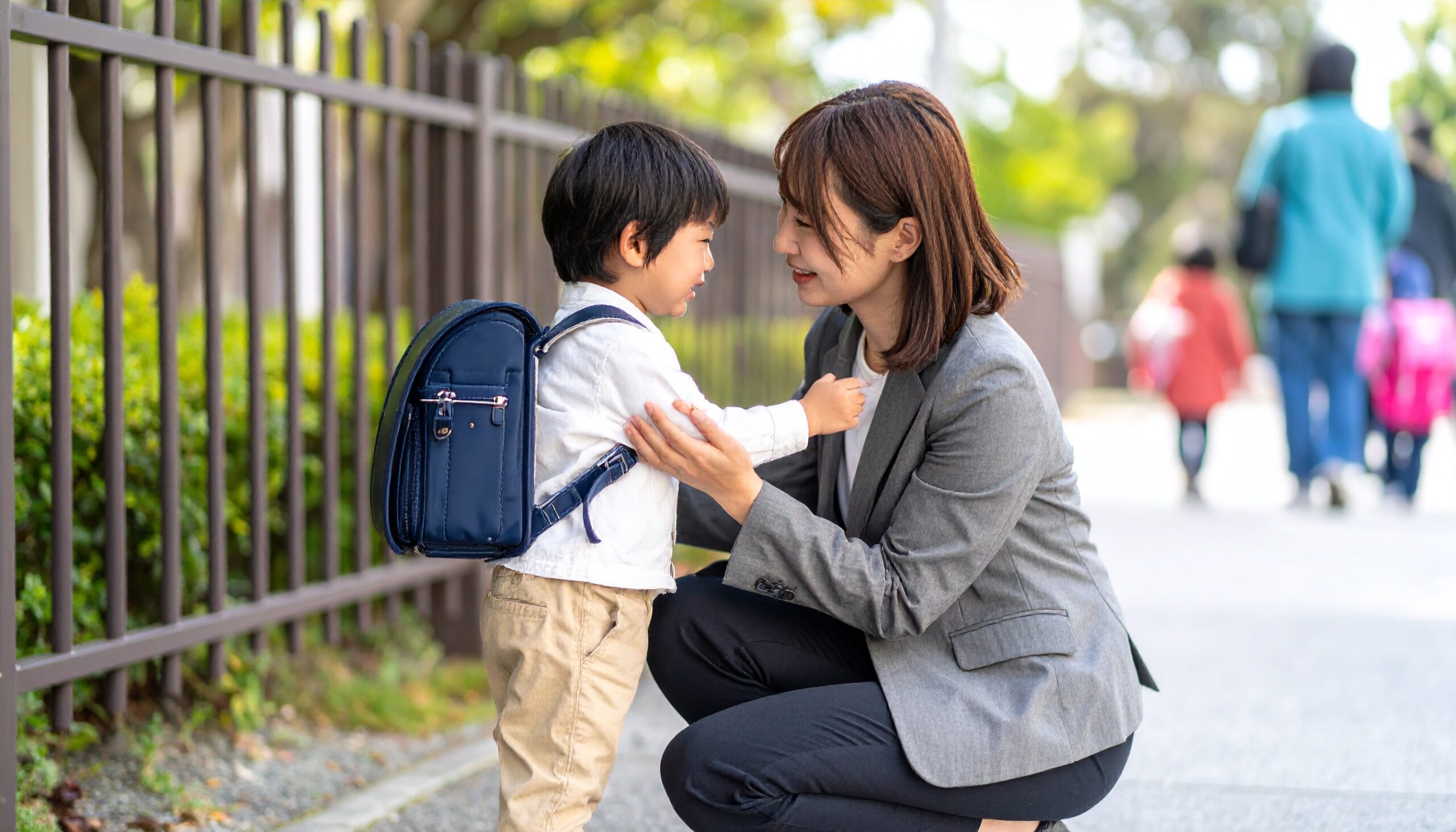 <img src="画像URL" alt="保育園の前で日本人の母親が泣いている3歳の男の子を優しくなだめている様子。" />
