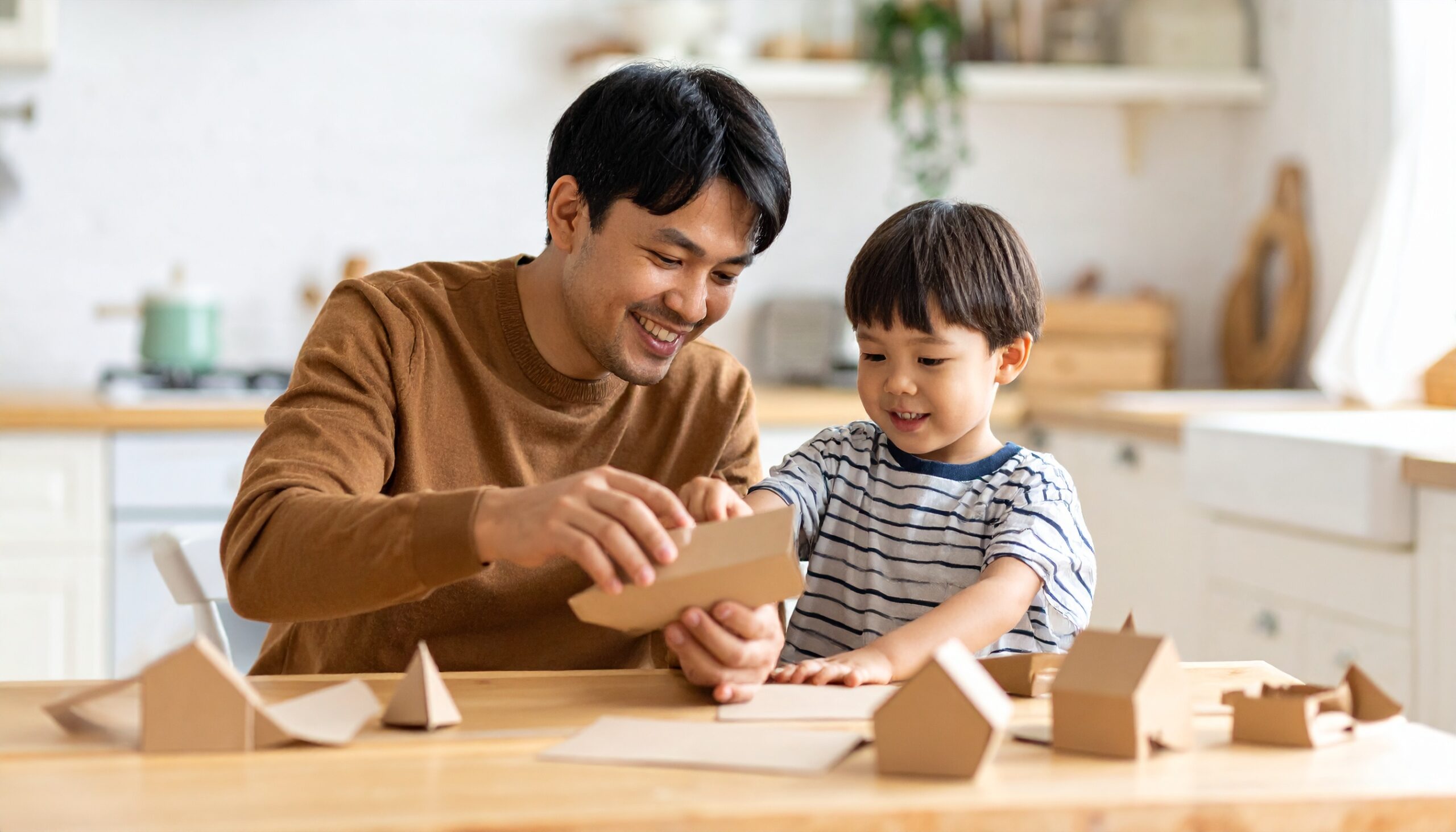 alt="キッチンのテーブルで段ボール工作を楽しむ日本人の父親と3歳の息子。明るい自然光の中、カジュアルな服装で笑顔を見せる親子"

